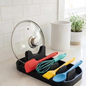 May include: A black kitchen utensil holder with a clear glass lid, red spatula, teal whisk, yellow brush, and blue spoon. The holder is on a white countertop with a white tile backsplash.