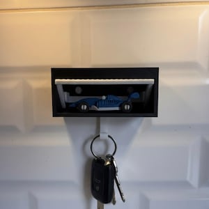 May include: A black key holder with a miniature blue race car displayed inside a small garage. The key holder has a white hook with a set of keys hanging from it. The key holder is mounted on a white tiled wall.