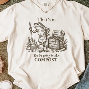 May include: Ivory-colored t-shirt featuring a humorous illustration of a frog in an apron holding a pitchfork, with the text "That's it. You're going in the COMPOST." The shirt has a crew neck and short sleeves. A necklace is visible.