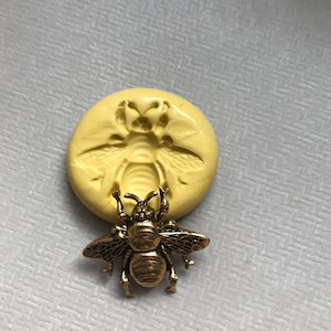 May include: A gold-colored bee charm with detailed wings and a textured body. The charm is sitting on a yellow silicone mold of a bee.