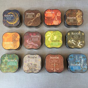 May include: Twelve ink pads in a variety of colors and designs. Each pad has a square shape with rounded corners and features a decorative design with a vintage aesthetic. The ink pads are labeled "StazOn" and include colors like Midnight Blue, Spiced Chai, Castle Poppy, Orange Zest, Emerald City, Cloudy Sky, Claret, and Blue Hawaii.
