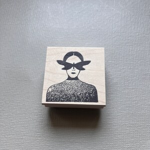 May include: A wooden stamp with a black and white illustration of a person with a butterfly covering their eyes.