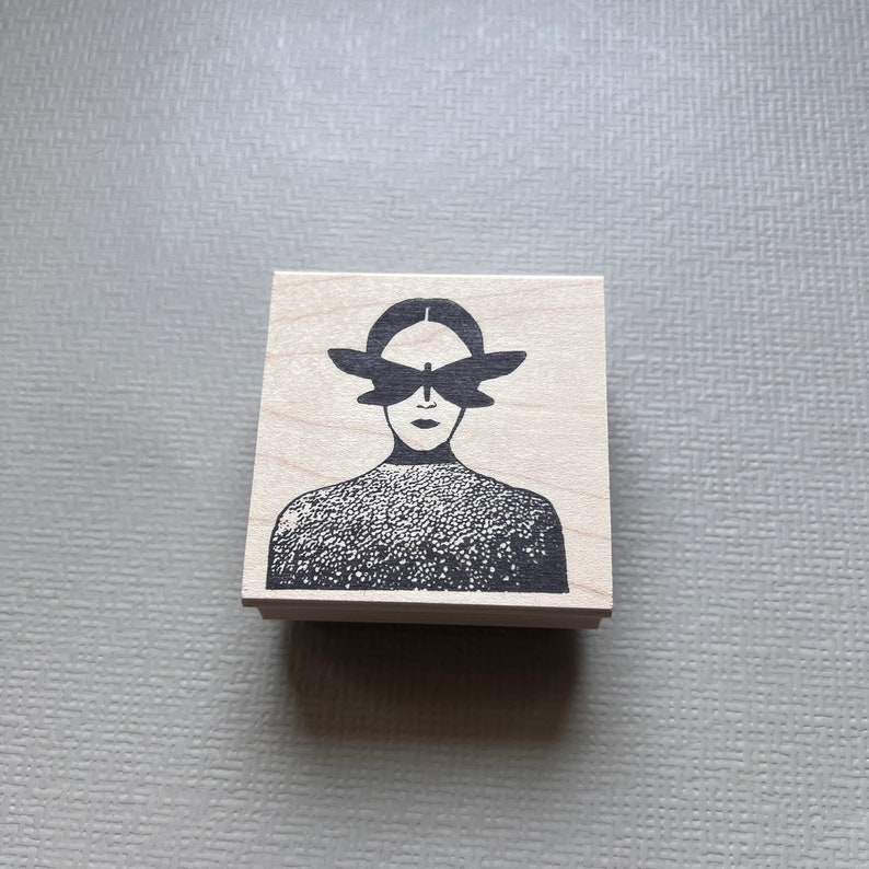 May include: A wooden stamp with a black and white illustration of a person with a butterfly covering their eyes.