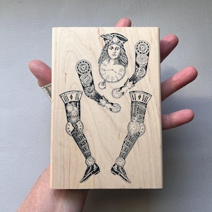 May include: A wooden stamp featuring a steampunk-style design. The design includes a woman's face with a clock, clockwork arms, and clockwork legs. The stamp is held in a hand.