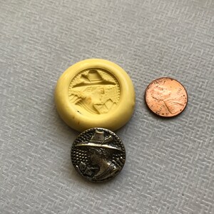 May include: A silver metal button with a raised design of a person wearing a hat and holding a bow and arrow. The button is about the size of a US penny.