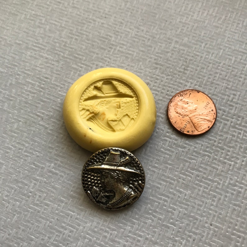 May include: A silver metal button with a raised design of a person wearing a hat and holding a bow and arrow. The button is about the size of a US penny.