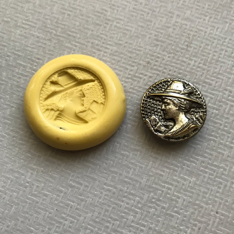 May include: A close-up of a vintage metal button featuring a detailed profile of a person wearing a hat. The button is silver in color and has a textured design.
