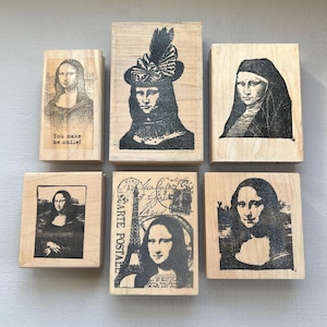 May include: A set of six rectangular wooden rubber stamps featuring black ink illustrations of the Mona Lisa. One stamp includes the text 'You make me smile!' and another features the Eiffel Tower with the text 'CARTE POSTALE'.