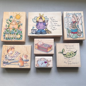May include: A collection of wooden rubber stamps with various illustrations. Designs include a cat with flowers, a girl with a broom, a boy fishing, mice with toothpaste, a dog in a jar, and peas in a pod. Text includes "Warm Thoughts", "Best Buds Forever!"