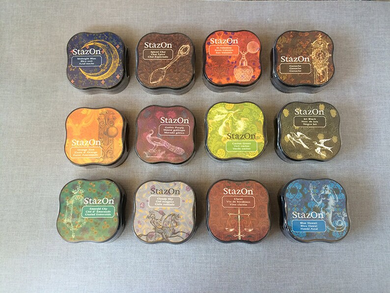 May include: Twelve ink pads in a variety of colors and designs. Each pad has a square shape with rounded corners and features a decorative design with a vintage aesthetic. The ink pads are labeled "StazOn" and include colors like Midnight Blue, Spiced Chai, Castle Poppy, Orange Zest, Emerald City, Cloudy Sky, Claret, and Blue Hawaii.