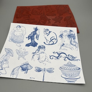 May include: A rectangular red rubber stamp sheet featuring various embossed designs, accompanied by a white paper printout displaying blue ink illustrations of a sumo wrestler, a person in traditional attire, a cat, a dragon, and a lidded bowl.