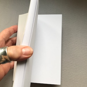 May include: A white blank notebook with a white cover and a white spine. The notebook is open to a blank page.