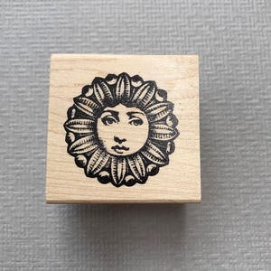 May include: A wooden stamp with a black and white illustration of a sun with a face. The sun has a floral design around it.