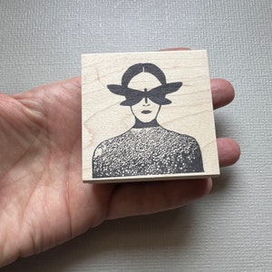 May include: A wooden block stamp with a black and white illustration of a person with a butterfly over their eyes.