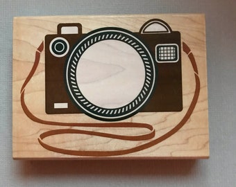 May include: A wooden stamp with a brown camera design, a white centre, and a brown strap. The camera has a circular lens and a rectangular viewfinder. The stamp is on a light-coloured wooden block.