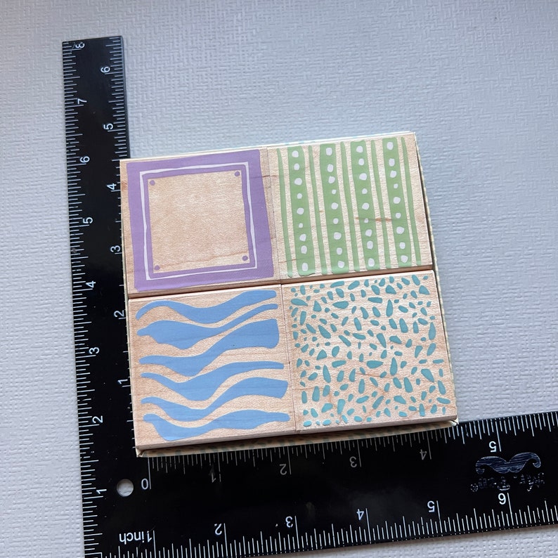 May include: Four wooden blocks with different patterns. The top left block has a purple border and a blank space inside. The top right block has green and white stripes. The bottom left block has blue wavy lines. The bottom right block has a light blue and green speckled pattern.