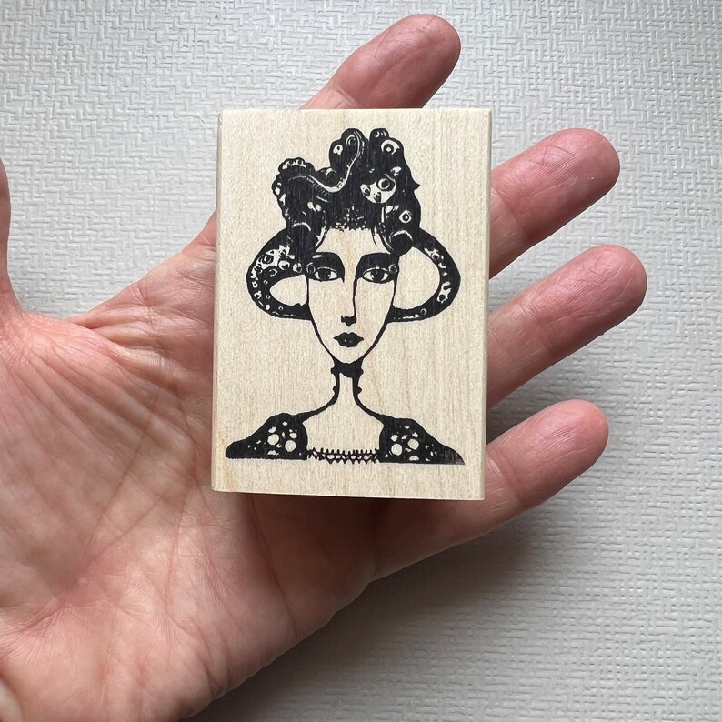 May include: A black and white rubber stamp featuring a woman with an octopus for hair. The woman is wearing a patterned dress and has a neutral expression.