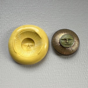 May include: A circular yellow silicone mold featuring a recessed sun face design with radiating lines, positioned next to a round metallic button with a bronze-toned sunburst border and a central pale green face with defined facial features.