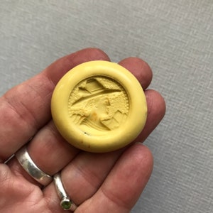 May include: A yellow round wax seal with a raised design of a person wearing a hat and a patterned garment.