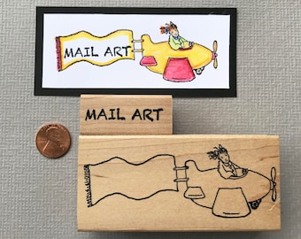 May include: Two wooden rubber stamps with a mail art theme. The first stamp features a yellow airplane with a red banner that says "MAIL ART". The second stamp features a black and white illustration of an airplane with a banner.