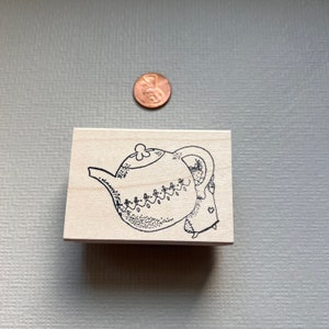 May include: A wooden stamp with a black and white illustration of a teapot with a person inside. The teapot has a floral design and the person is wearing a dress.