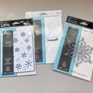 May include: Three Memory Box craft die packages. One features a snowflake design, another a snow globe, and the third a snowflake. The packages are clear with black and teal accents. The text "100% Steel Craft Die" is visible.