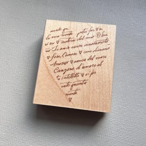 May include: A rectangular wooden stamp featuring a heart-shaped arrangement of Italian text in a dark brown cursive script. The text includes phrases like 'la mia mente', 'materia del mio', and 'Canzone d'amore', interspersed with small heart icons.
