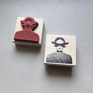 May include: Two wooden rubber stamps with black ink. The first stamp features a person with a red face and a black hat. The second stamp features a person with a black and white face and a black butterfly design over their eyes.