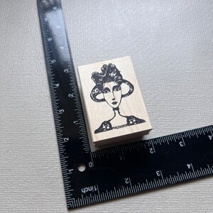 May include: A wooden rubber stamp with a black and white illustration of a woman with a large, ornate hat. The woman has a long neck and a simple dress.