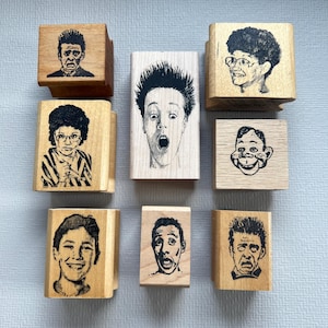 May include: A collection of wooden stamps featuring expressive faces. Each stamp displays a different sketched portrait, showcasing a range of emotions from surprise to fear. The stamps are made of light-colored wood and are arranged on a light blue surface.