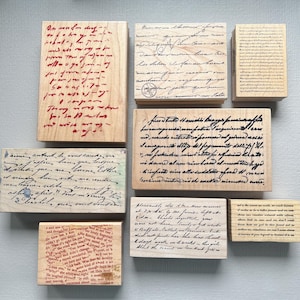 May include: A collection of wooden stamps with handwritten script in various fonts and ink colors. The stamps feature text in red, black, and blue inks. The stamps are rectangular and square, ideal for scrapbooking or journaling.