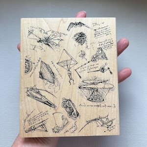 May include: A rectangular light-colored wooden block featuring black ink illustrations of various Leonardo da Vinci inventions, including a flying machine, a helicopter, and mechanical sketches, accompanied by handwritten-style notes.