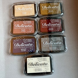 May include: Eight ink pads in clear plastic containers with black lids. Each pad has a different color ink and features the text "Rich and Radiant Riche et Radiant Rico y Radiante Delicata Pigment Ink/Encre Pigmentée/Tinta con Pigmento" and the name of the color in English, French, and Spanish.
