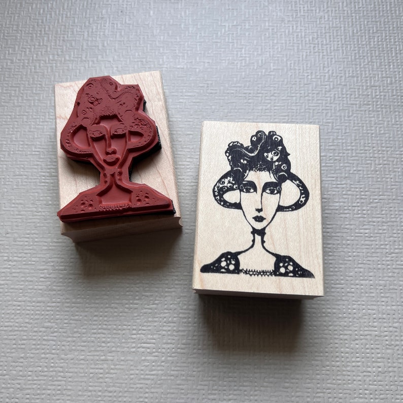 May include: Two wooden mounted rubber stamps featuring black and white illustrations of women with elaborate hairstyles. The stamps are on a light beige surface.
