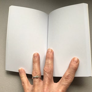 May include: A blank, white notebook with a soft, white cover is open to a blank page. The notebook is lying flat on a light grey surface.