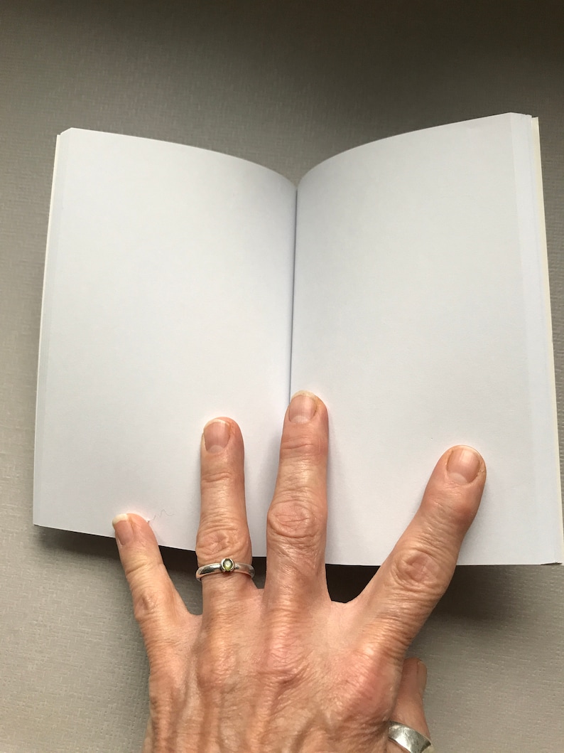 May include: A blank, white notebook with a soft, white cover is open to a blank page. The notebook is lying flat on a light grey surface.