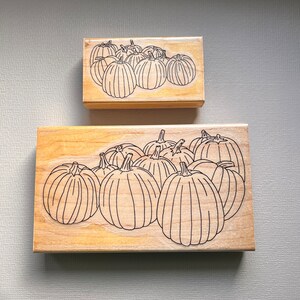 May include: Two wooden stamps with a pumpkin design. The larger stamp features a group of pumpkins, while the smaller stamp has a similar design. The pumpkins are outlined in black on a light wood background.