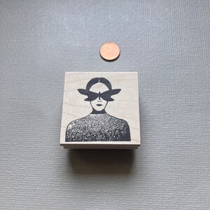 May include: A wooden stamp with a black and white illustration of a person with a butterfly covering their eyes.
