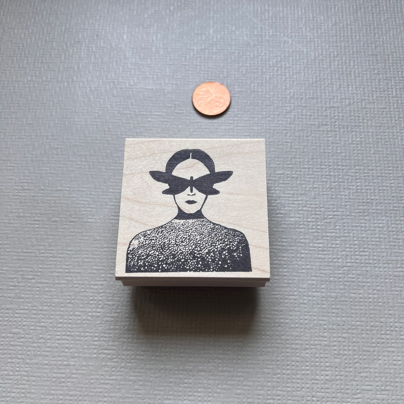 May include: A wooden stamp with a black and white illustration of a person with a butterfly covering their eyes.
