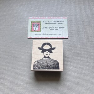 May include: A wooden rubber stamp featuring a black and white illustration of a person with butterfly wings over their eyes. The person is wearing a textured sweater.