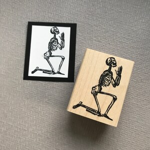 May include: A wooden rubber stamp with a black and white image of a skeleton kneeling with hands raised in prayer.