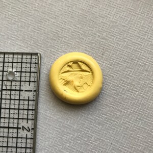 May include: A yellow round button with a raised image of a person wearing a hat and a coat. The person is facing to the right and has a beard.