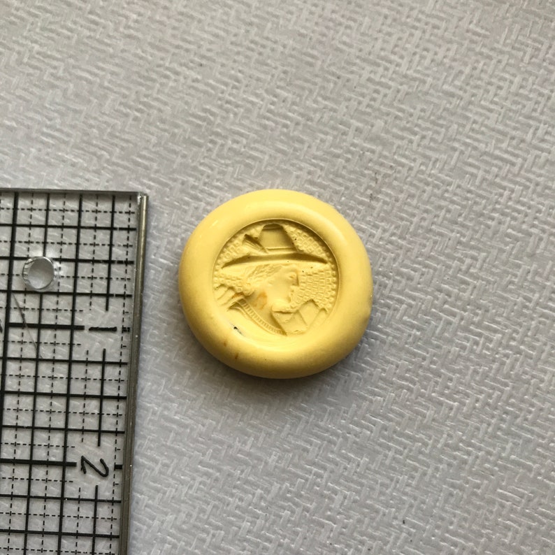 May include: A yellow round button with a raised image of a person wearing a hat and a coat. The person is facing to the right and has a beard.