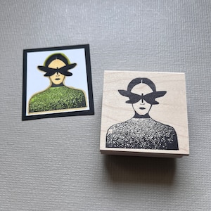 May include: A wooden rubber stamp with a black and white design of a person with a butterfly over their eyes. The person is wearing a textured sweater.