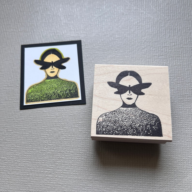 May include: A wooden rubber stamp with a black and white design of a person with a butterfly over their eyes. The person is wearing a textured sweater.