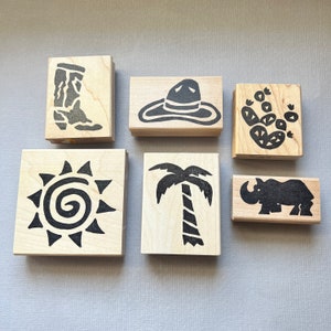 May include: A collection of six wooden stamps with black ink designs. The stamps feature a cowboy boot, a sombrero, a cactus, a sun, a palm tree, and a rhinoceros. The stamps are arranged on a light gray surface.