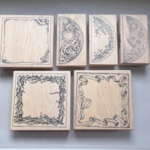 May include: A collection of six wooden stamps with various black ink designs. The stamps feature frames, ribbons, and botanical elements. Some stamps are square, while others are semi-circular. The designs are detailed and suitable for crafting.