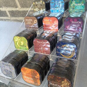 May include: A display of StazOn ink pads in various colors and designs. The ink pads are arranged in a clear acrylic display stand. The ink pads are labeled with the brand name "StazOn" and the color or design of the ink.