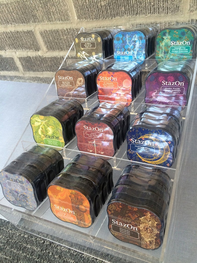 May include: A display of StazOn ink pads in various colors and designs. The ink pads are arranged in a clear acrylic display stand. The ink pads are labeled with the brand name "StazOn" and the color or design of the ink.
