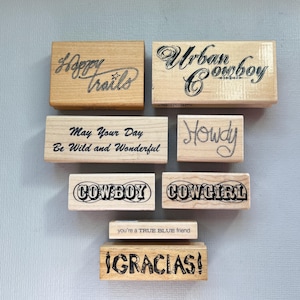May include: A collection of eight rectangular wooden rubber stamps featuring black text and designs. The stamps include phrases such as Happy trails, Urban Cowboy, May Your Day Be Wild and Wonderful, Howdy, COWBOY, COWGIRL, you're a TRUE BLUE friend, and GRACIAS.